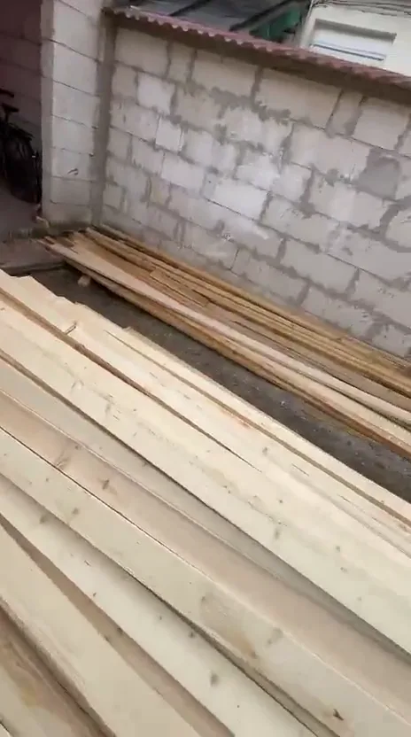 Grinzi și căpriori de brad proaspăt tăiați, stivuiți lângă zidul unei case, înainte de montaj.