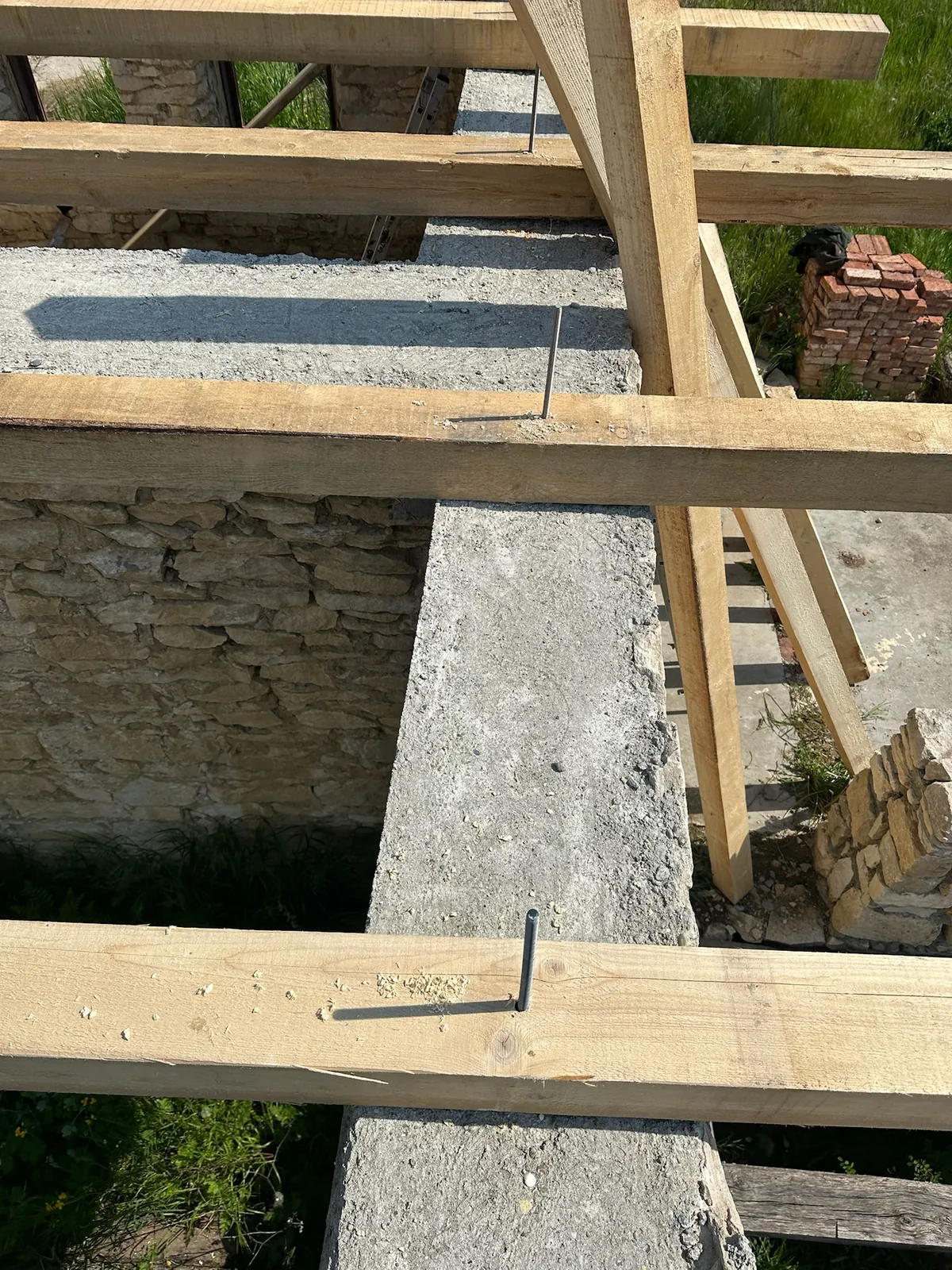 Grinzi noi de lemn fixate cu ancore metalice în centura de beton a unei case de piatră, cu stiva de cărămizi în fundal.