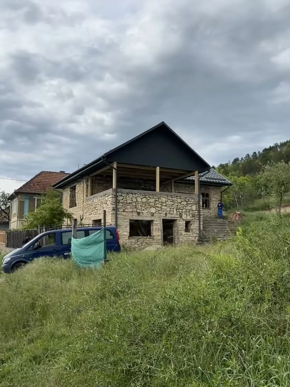 Casă de piatră cu etajul mansardat recent, cu acoperiș negru nou și terasă de lemn, pe un teren cu iarbă înaltă.