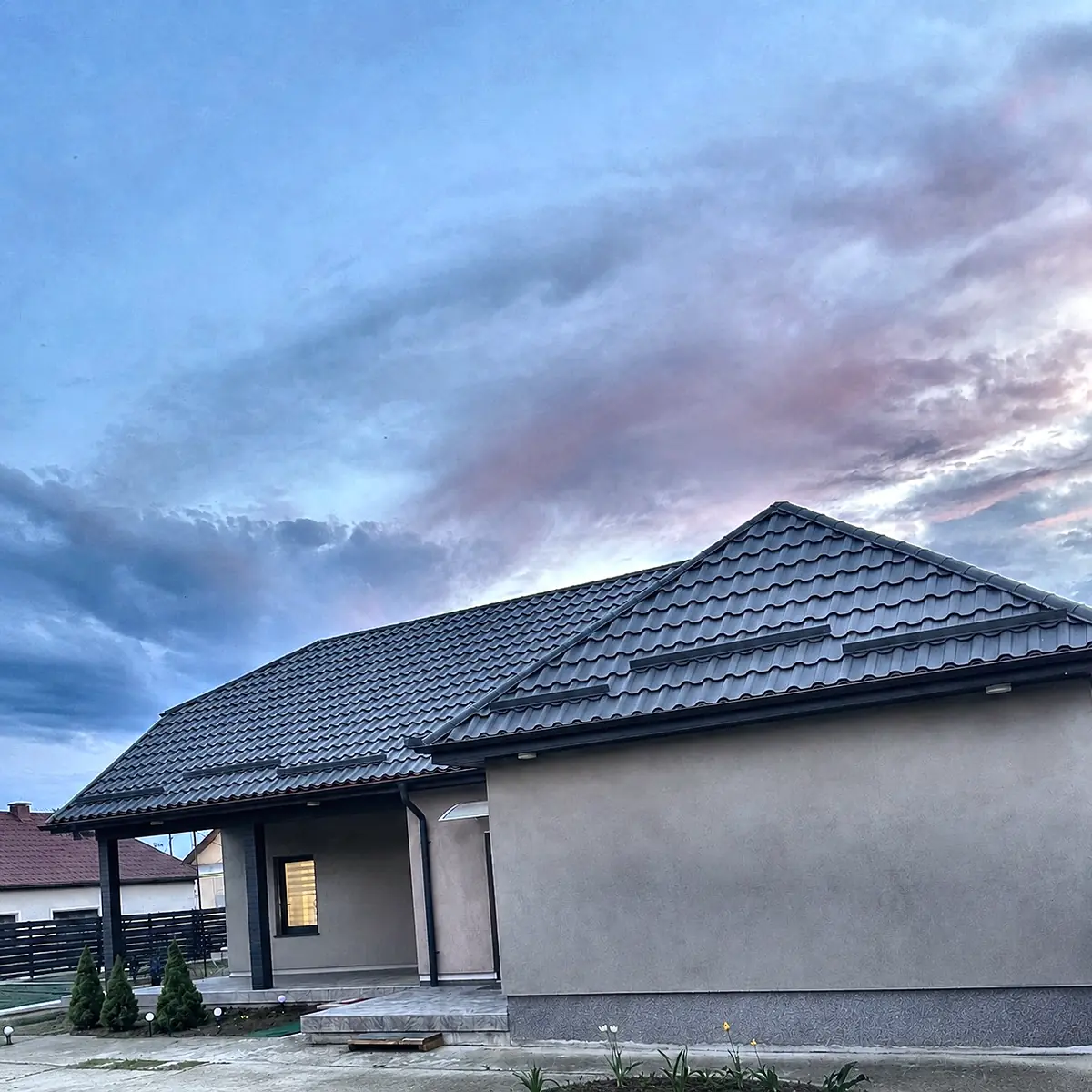 Casă nouă cu acoperiș de țiglă metalică antracit, fotografiată în amurg dinspre curte.