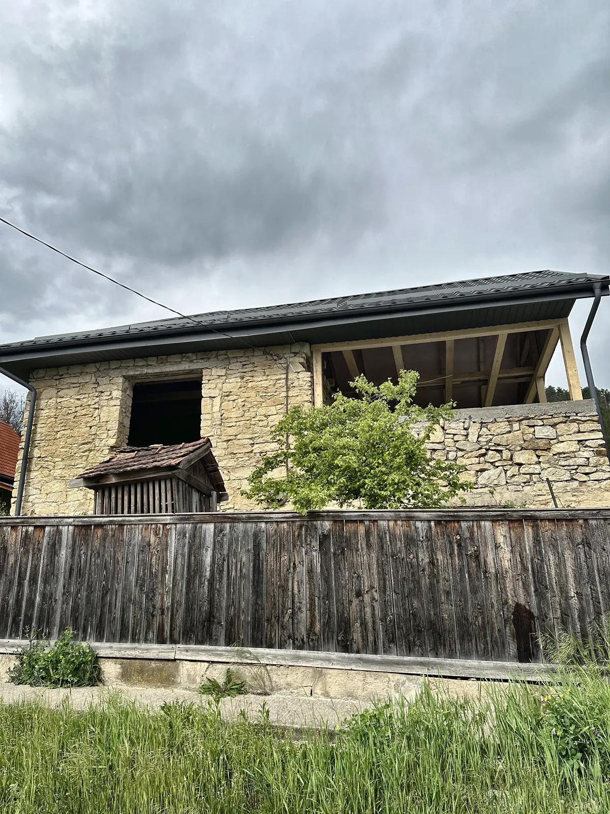 Casă de piatră cu acoperișul vechi încă pe ea, văzută de peste gardul de scândură.
