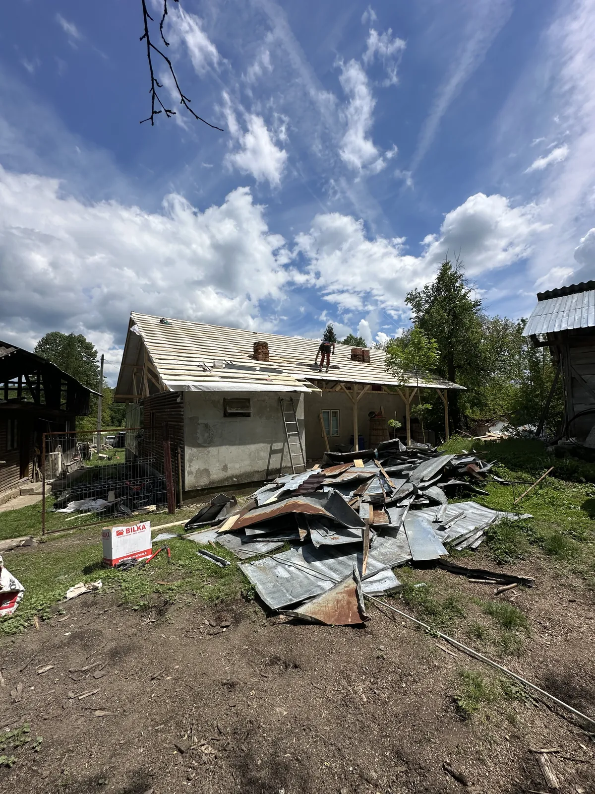 Curte de șantier cu foile de tablă veche demontate și strânse grămadă, iar în fundal casa cu astereala nouă montată pe acoperiș.