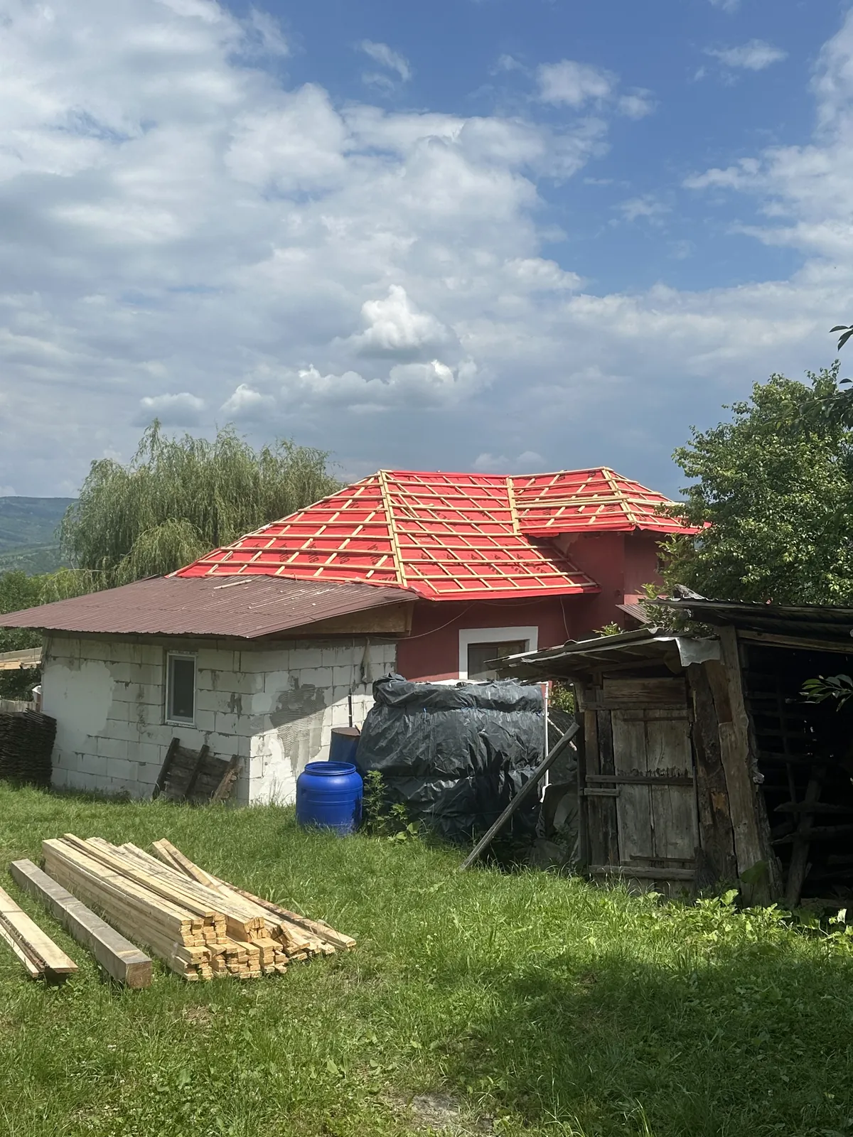 Casă roșie cu acoperișul în refacere: folie roșie și șipci proaspăt montate pe șarpantă, sub cer senin de vară.