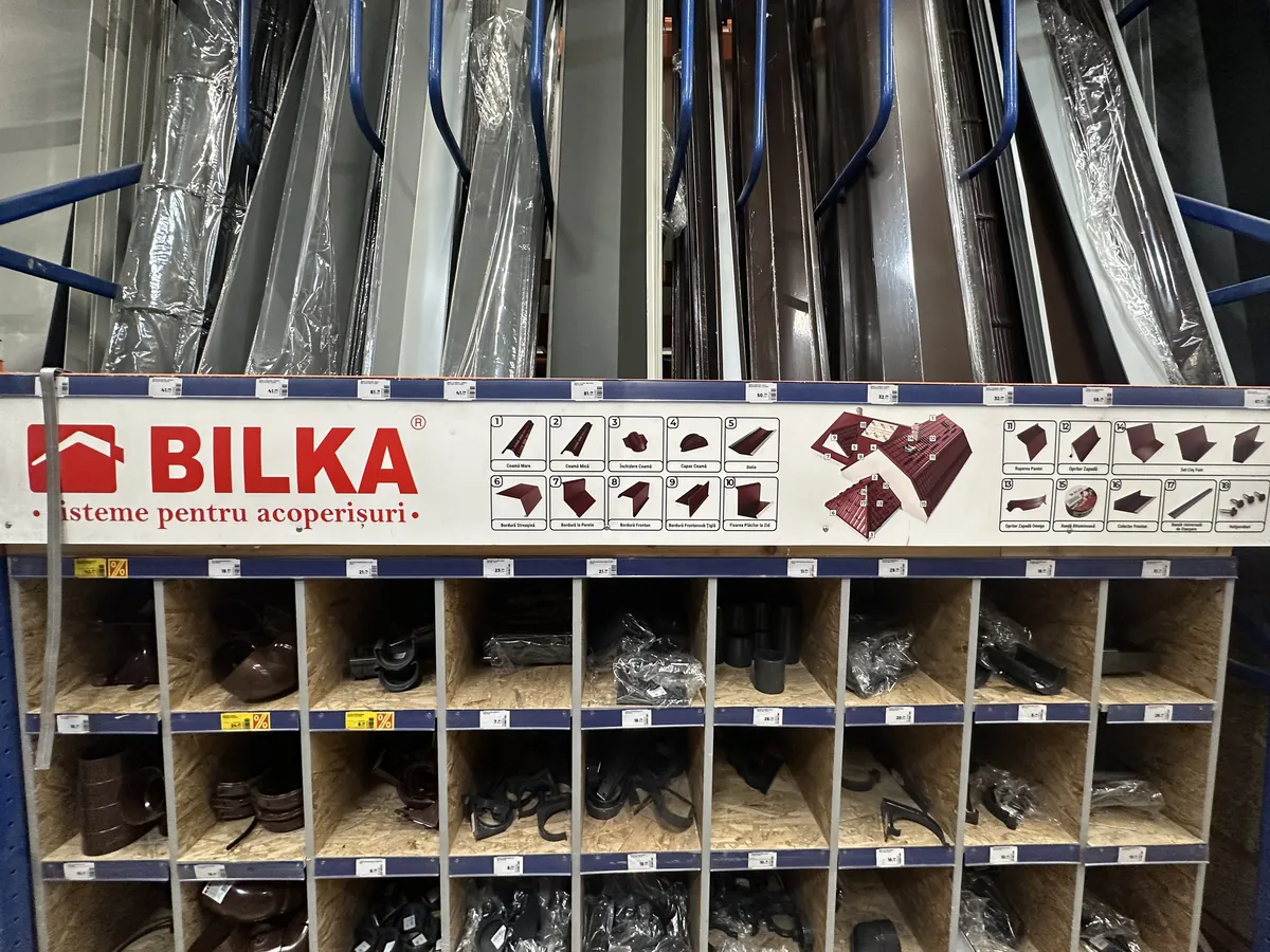 Raft de accesorii BILKA într-un depozit: coame, dolii, opritoare de zăpadă și cleme, pe compartimente etichetate.