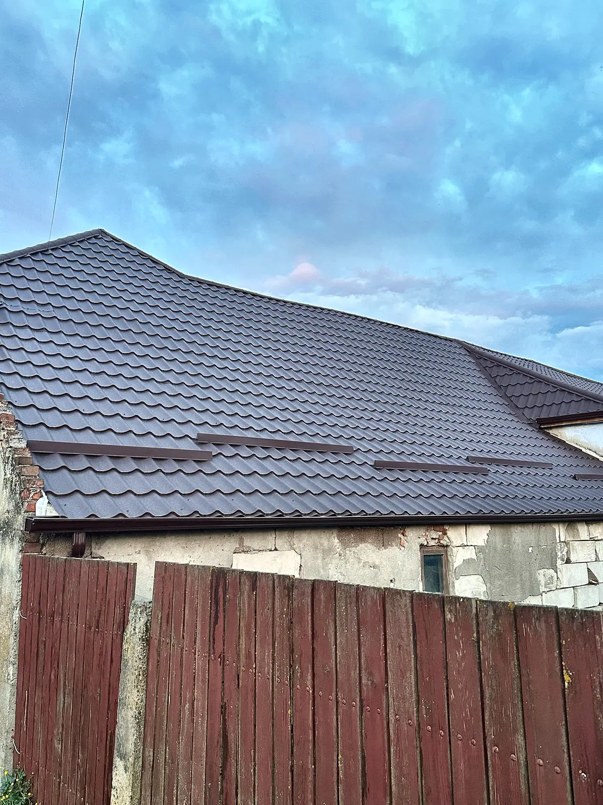 Acoperiș de țiglă metalică maro, văzut peste un gard de lemn roșu.