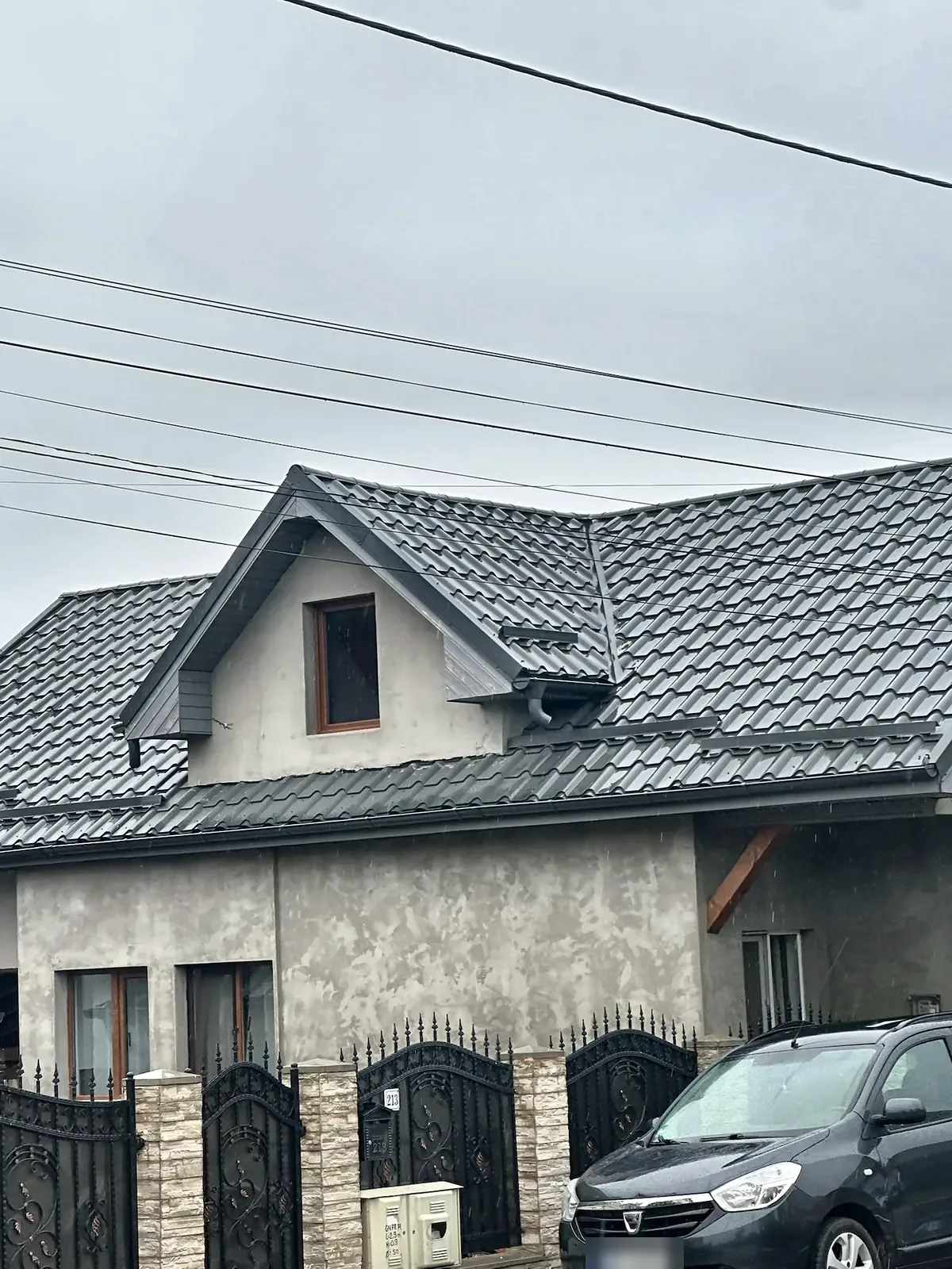 Casă cu acoperiș de țiglă metalică gri și lucarnă, văzută de la poarta de fier.